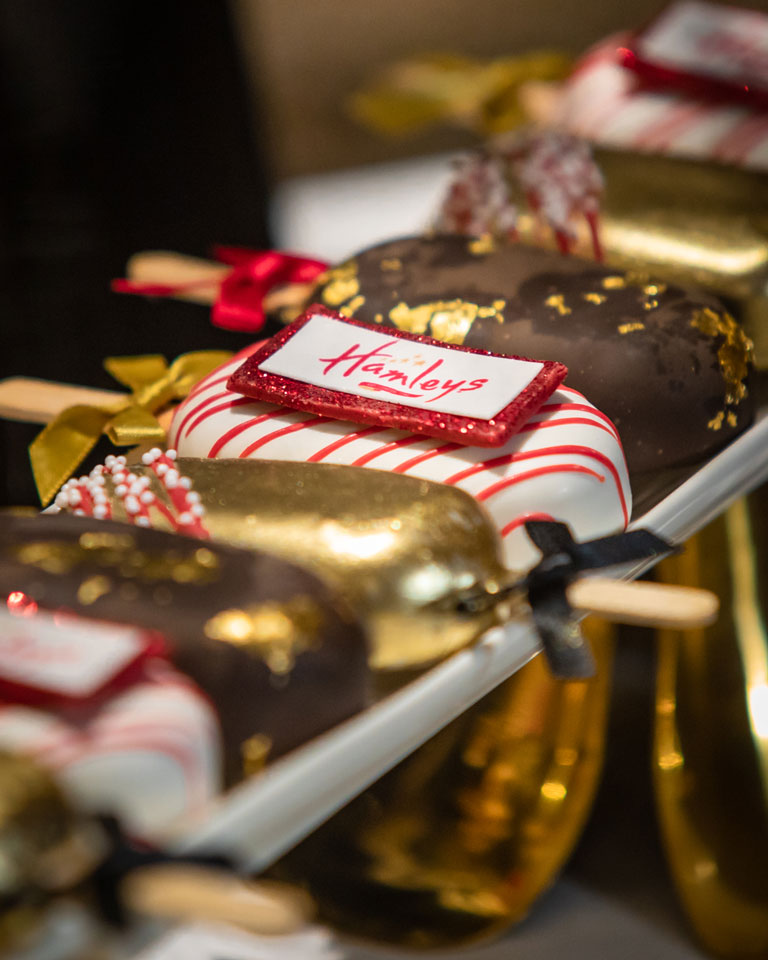 Lolli cakes in chocolate red and gold colours with Hamleys logo tied with ribbon around the lolli sticks